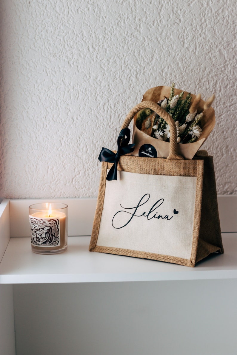 May include: A brown burlap tote bag with a white canvas insert. The bag has a black ribbon bow and the name "Selina" written in black script on the front. The bag is filled with a bouquet of dried flowers and a small brown paper bag with a "Handmade" label.