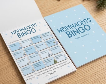 May include: A light blue Christmas bingo game pad with white snowflake patterns. The cover reads WEIHNACHTS BINGO FROHE WEIHNACHTEN. The open page features a grid of squares with German text, instructions, and small holiday illustrations.