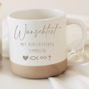 Può includere: Una tazza in ceramica bianca con finitura maculata e base beige. La tazza presenta il testo "Wunschtext" e "MIT VERSCHIEDENEN SYMBOLEN" in marrone chiaro, insieme a simboli di cuore, pesce, infinito e croce. La tazza ha un manico curvo.