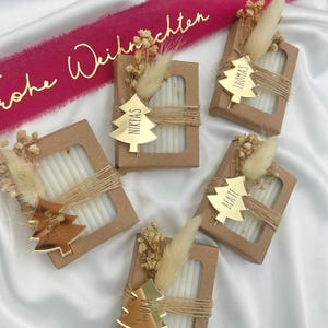 Può includere: Cinque piccole scatole regalo rettangolari con finestra, ciascuna contenente candele bianche. Le scatole sono avvolte con spago e decorate con un'etichetta dorata a forma di albero con un nome. Fiori secchi e un nastro con testo dorato completano il look festivo.