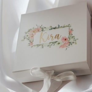 Op de afbeelding: Witte cadeaudoos met een bloemenontwerp en de naam "Kira" in gouden letters. De doos is vastgebonden met een wit satijnen lint.