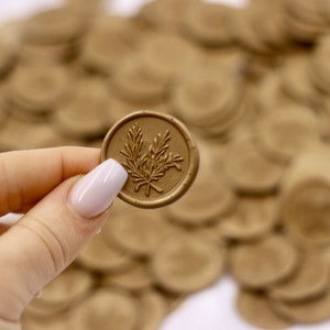 Op de afbeelding: Een close-up van een gouden waszegel met een takje bladeren ontwerp. De zegel wordt vastgehouden door een hand met witte nagellak. De zegel wordt omringd door een stapel andere gouden waszegels.