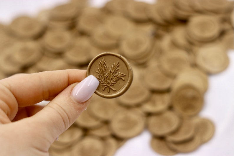 Op de afbeelding: Een close-up van een gouden waszegel met een takje bladeren ontwerp. De zegel wordt vastgehouden door een hand met witte nagellak. De zegel wordt omringd door een stapel andere gouden waszegels.