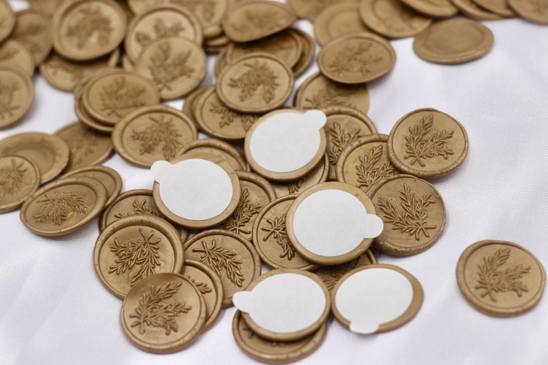 Op de afbeelding: Een stapel gouden waszegels met een bladerenontwerp. Sommige zegels hebben witte zelfklevende etiketten op de achterkant.