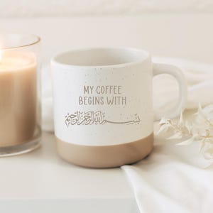 Könnte beinhalten: Ein zweifarbiger Keramikbecher mit einem gesprenkelten weißen oberen Teil und einem einfarbigen beigen Boden. Der Becher ist mit dem Text "MY COFFEE BEGINS WITH" in einer dunkelgrauen serifenlosen Schrift und darunter arabischer Kalligrafie bedruckt.