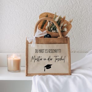 Op de afbeelding: Een afstudeercadeauset met een jute draagtas met de tekst "DU HAST ES GESCHAFFT! Master in der Tasche!" en een afstudeerpet-afbeelding. De tas bevat gedroogde bloemen en staat naast een brandende kaars.
