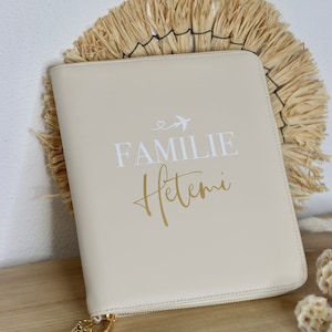 Pode incluir: Uma bolsa de viagem bege com zíper, com um zíper dourado e uma alça branca. A bolsa tem o texto "FAMILIE Hetemi" impresso nela em letras brancas e douradas.