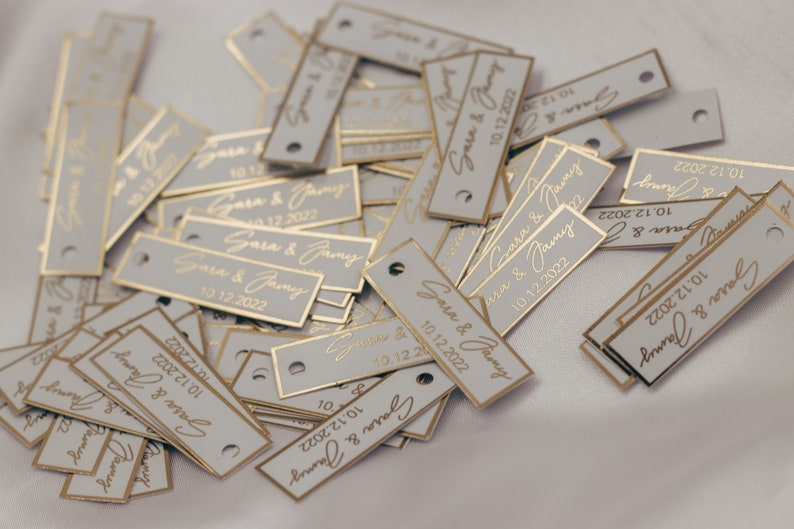 May include: A pile of small, rectangular, gray tags with gold foil lettering. The tags have the text "Sara & Jamy" and the date "10.12.2022" printed on them.