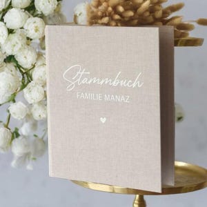 Könnte beinhalten: Ein beigefarbenes, mit Leinen bezogenes Buch mit dem Text "Stammbuch FAMILIE MANAZ" in weißen Buchstaben. Ein kleines weißes Herz befindet sich mittig unter dem Text.