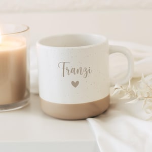 Op de afbeelding: Witte keramische mok met een gespikkelde afwerking en een bruine basis. De mok heeft de naam "Franzi" in een scriptlettertype, met een klein hartsymbool. Een brandende kaars in een glazen pot is zichtbaar op de achtergrond.