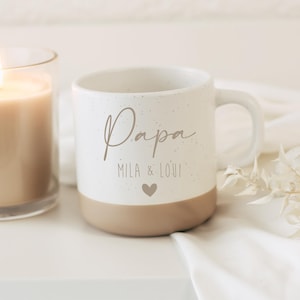 Pode incluir: Uma caneca de cer&acirc;mica branca com acabamento salpicado e base bege. A caneca apresenta a palavra "Papa" em escrita cursiva, juntamente com "MILA & LOUI" e um s&iacute;mbolo de cora&ccedil;&atilde;o. Uma vela acesa e tecido branco no fundo.