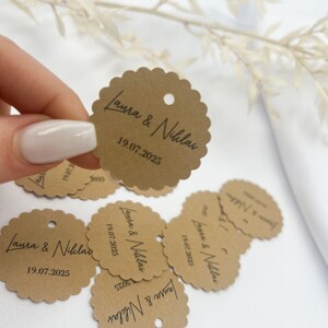 May include: Brown paper circle tags with scalloped edges and a hole punched at the top. The tags have the text "Laura & Niklas" and the date "19.07.2025" printed in black.