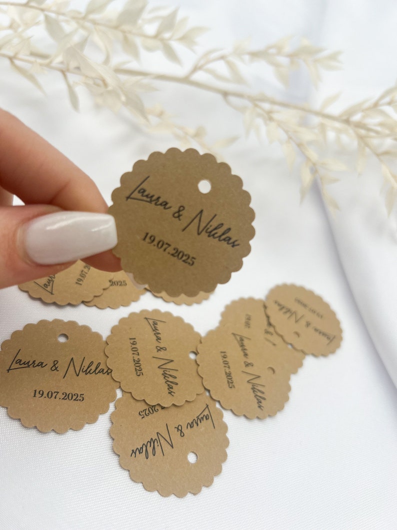 May include: Brown paper circle tags with scalloped edges and a hole punched at the top. The tags have the text "Laura & Niklas" and the date "19.07.2025" printed in black.