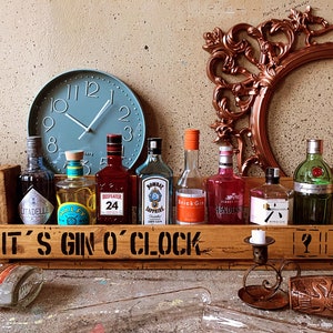 Könnte beinhalten: Eine Holzkiste mit dem Text "It's Gin O'Clock" auf der Vorderseite. Die Kiste ist gefüllt mit verschiedenen Gin-Flaschen, darunter Bombay Sapphire, Hendricks und Tanqueray. Hinter der Kiste hängt auch eine Uhr an der Wand.