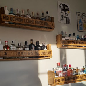 May include: Three wooden shelves with a variety of gin bottles. The shelves are decorated with the text "Gib deinem Leben einen Gin", "Nach meinem Gin", "Der Gin des Lebens", and "Und am Ende ergibt alles einen Gin".
