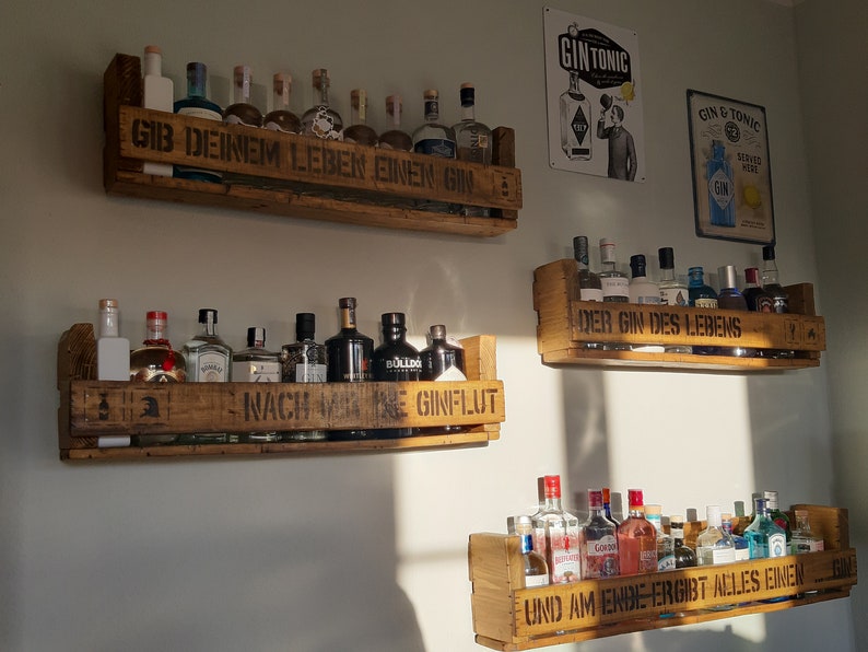 May include: Three wooden shelves with a variety of gin bottles. The shelves are decorated with the text "Gib deinem Leben einen Gin", "Nach meinem Gin", "Der Gin des Lebens", and "Und am Ende ergibt alles einen Gin".