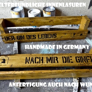 May include: Two wooden shelves with the text "Der Gin des Lebens" and "Handmade in Germany" on the top shelf and "Wach mir die Gin-Flut" on the bottom shelf. The shelves are stained a light brown color and have a rustic appearance.