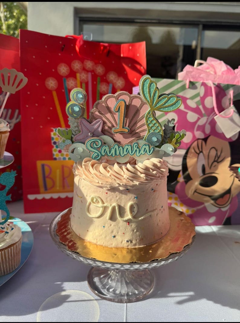 Puede incluir: Un pastel glaseado de color rosa y blanco con un adorno de tem&aacute;tica de sirena. El adorno presenta una concha, una cola de sirena y el n&uacute;mero 1. El pastel est&aacute; decorado con chispas de colores y la palabra "uno" est&aacute; escrita en glaseado blanco. El pastel est&aacute; sobre un soporte para pastel de vidrio transparente.