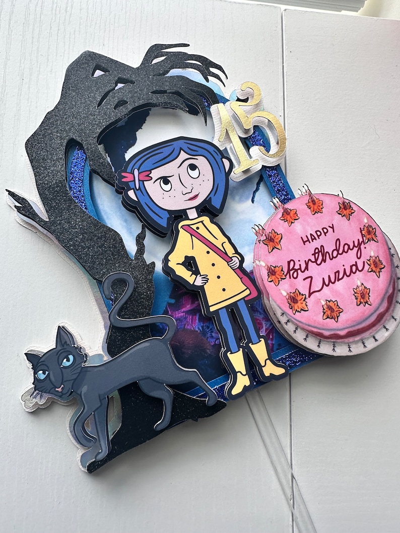 May include: A cake topper featuring a black silhouette of a monster, a girl with blue hair wearing a yellow raincoat, a black cat, and a pink cake with the text "Happy Birthday Zuzia".