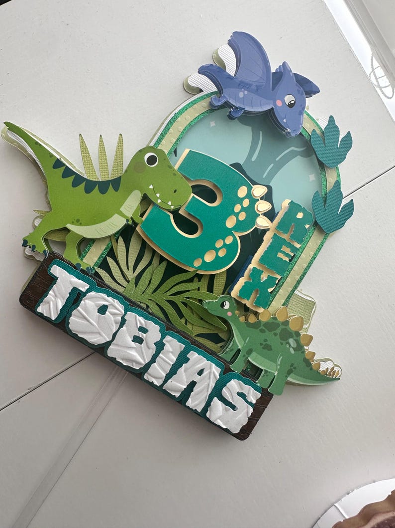 Puede incluir: Topper de pastel de dinosaurio verde y azul con el n&uacute;mero 3 y el nombre Tobias. El topper presenta un dinosaurio verde, un pterod&aacute;ctilo azul y hojas verdes.