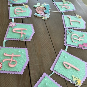 Puede incluir: Una pancarta de cumplea&ntilde;os con tem&aacute;tica de sirena con las letras "Happy Birthday" en rosa sobre un fondo verde y morado. Las letras est&aacute;n decoradas con purpurina y lentejuelas.