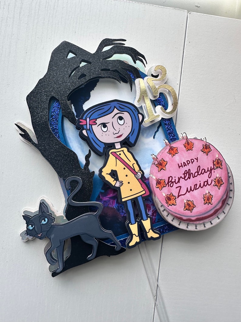 May include: A cake topper featuring a black cat, a girl with blue hair, a yellow raincoat, and a black silhouette of a tree. The cake topper also includes a pink cake with the text "Happy Birthday Zuzu" and the number "13" in gold.
