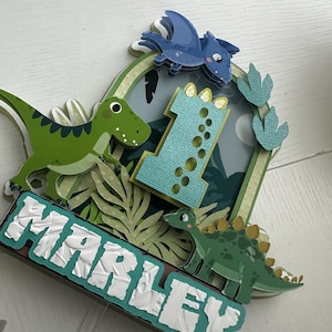 Puede incluir: Un adorno para pastel de dinosaurio verde y azul con el nombre "Marley" y el n&uacute;mero "1" en &eacute;l. El adorno presenta un dinosaurio verde, un pterod&aacute;ctilo azul y huellas de dinosaurio verdes.