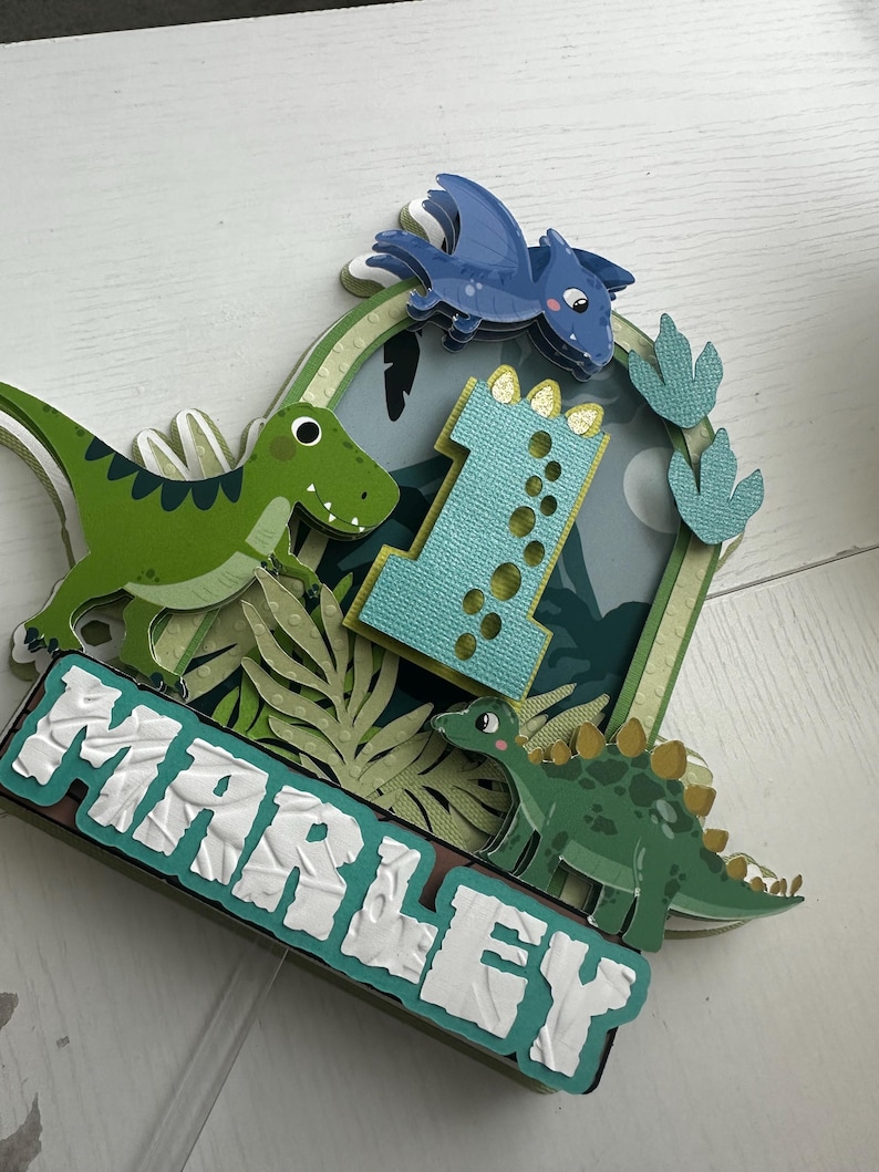 Puede incluir: Un adorno para pastel de dinosaurio verde y azul con el nombre "Marley" y el n&uacute;mero "1" en &eacute;l. El adorno presenta un dinosaurio verde, un pterod&aacute;ctilo azul y huellas de dinosaurio verdes.