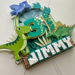Puede incluir: Topper de pastel de dinosaurio verde, azul y dorado con el nombre "JIMMY" y el n&uacute;mero "3" en &eacute;l.