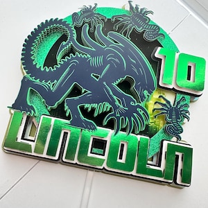 May include: A layered cake topper featuring a dark blue alien creature design with green glitter accents. The word "LINCOLN" and the number "10" are in green, with a white outline. The topper is likely for a birthday celebration.