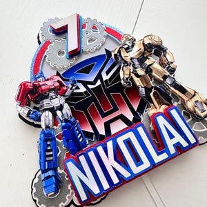 May include: A red, blue, and silver cake topper featuring the Transformers logo, Optimus Prime, Bumblebee, and the name "NIKOLAI".