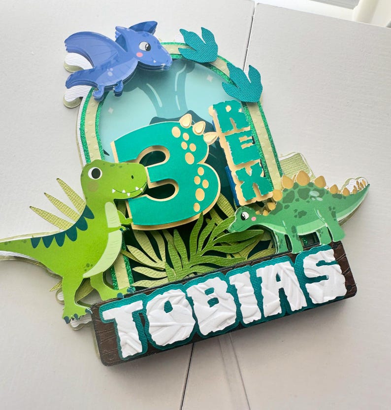 Puede incluir: Un adorno para pastel de dinosaurio verde y azul con el n&uacute;mero 3, el nombre Tobias y la palabra Rex. El adorno presenta un dinosaurio verde, un pterod&aacute;ctilo azul y hojas verdes.
