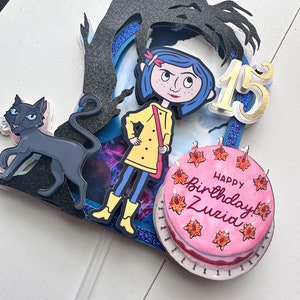 May include: A handmade cake topper featuring a girl with blue hair and a yellow raincoat, a black cat, a shadow with a hand, and a pink cake with the words "Happy Birthday! Zuzia" and the number "15".