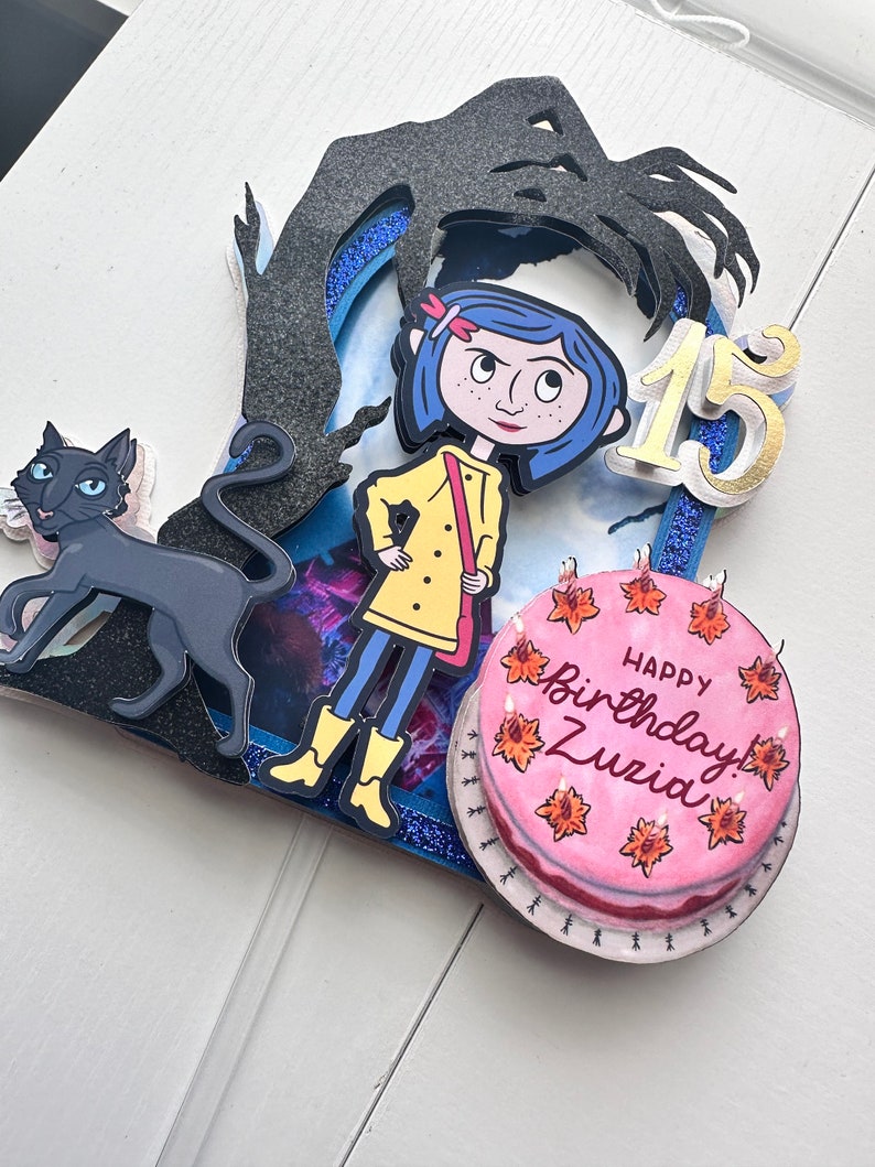 May include: A handmade cake topper featuring a girl with blue hair and a yellow raincoat, a black cat, a shadow with a hand, and a pink cake with the words "Happy Birthday! Zuzia" and the number "15".
