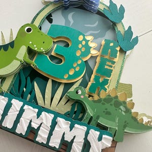Puede incluir: Un adorno para pastel de dinosaurio verde y dorado con un pterod&aacute;ctilo azul, un dinosaurio verde y el n&uacute;mero 3. El adorno tambi&eacute;n incluye el nombre "Jimmy" en letras blancas.