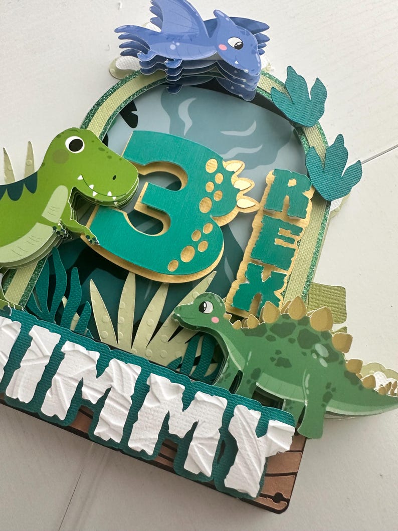 Puede incluir: Un adorno para pastel de dinosaurio verde y dorado con un pterod&aacute;ctilo azul, un dinosaurio verde y el n&uacute;mero 3. El adorno tambi&eacute;n incluye el nombre "Jimmy" en letras blancas.