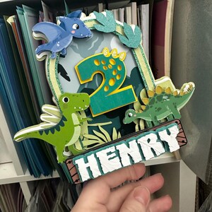 Puede incluir: Un adorno para pastel de dinosaurio verde y azul con el n&uacute;mero 2 y el nombre Henry. El adorno presenta un dinosaurio verde, un pterod&aacute;ctilo azul y un dinosaurio verde con manchas amarillas.