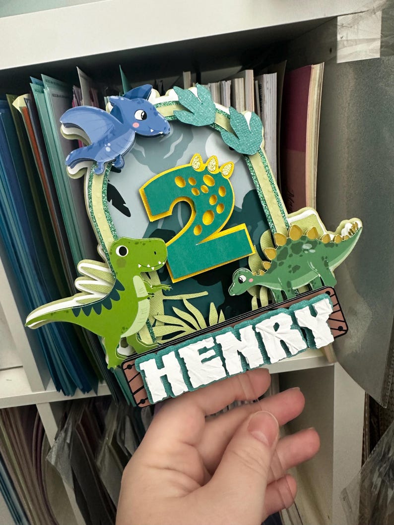 Puede incluir: Un adorno para pastel de dinosaurio verde y azul con el n&uacute;mero 2 y el nombre Henry. El adorno presenta un dinosaurio verde, un pterod&aacute;ctilo azul y un dinosaurio verde con manchas amarillas.