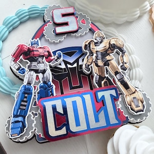 May include: A layered cake topper featuring the name COLT in blue and silver foil lettering, the number 5, and two robot figures. The design includes a central Autobot logo, grey gear shapes, and red accents, creating a multi-dimensional appearance.