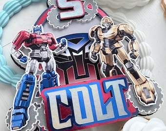 May include: A layered cake topper featuring the name COLT in blue and silver foil lettering, the number 5, and two robot figures. The design includes a central Autobot logo, grey gear shapes, and red accents, creating a multi-dimensional appearance.