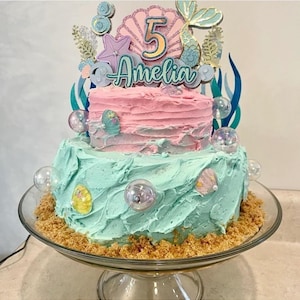 Puede incluir: Un pastel de dos pisos decorado con un tema de sirena. El piso superior es rosa con una cola de sirena, una concha y el n&uacute;mero 5. El piso inferior es azul claro con un borde marr&oacute;n arenoso. El pastel est&aacute; coronado con un letrero que dice "Amelia".