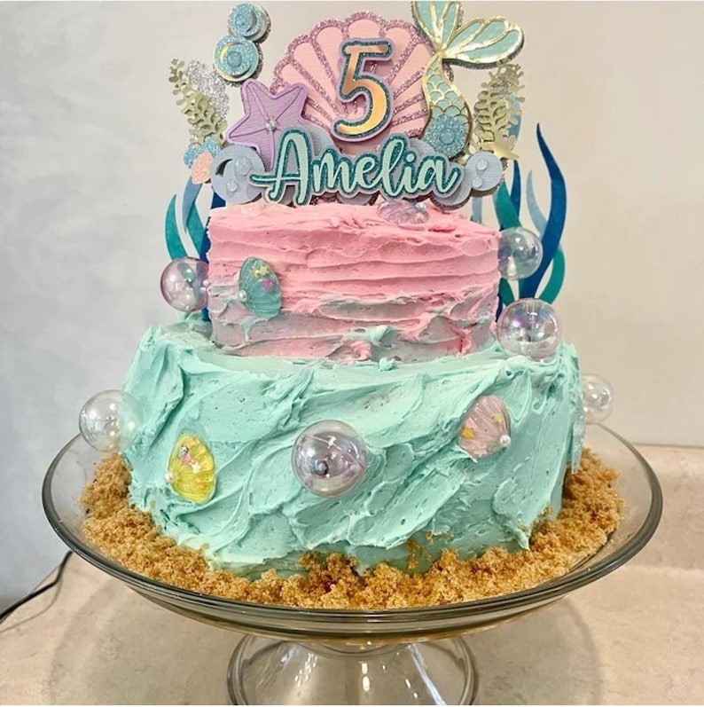 Puede incluir: Un pastel de dos pisos decorado con un tema de sirena. El piso superior es rosa con una cola de sirena, una concha y el n&uacute;mero 5. El piso inferior es azul claro con un borde marr&oacute;n arenoso. El pastel est&aacute; coronado con un letrero que dice "Amelia".