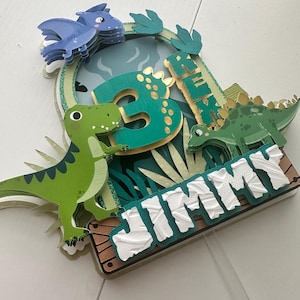 Puede incluir: Un adorno para pastel de dinosaurio verde y azul con el nombre "JIMMY" y el n&uacute;mero "3" en &eacute;l. El adorno presenta un dinosaurio verde, un dinosaurio azul y hojas verdes.