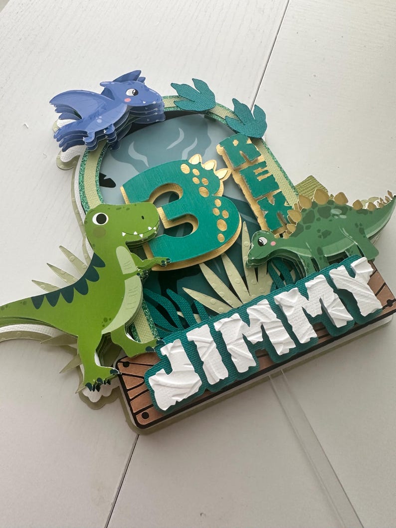 Puede incluir: Un adorno para pastel de dinosaurio verde y azul con el nombre "JIMMY" y el n&uacute;mero "3" en &eacute;l. El adorno presenta un dinosaurio verde, un dinosaurio azul y hojas verdes.