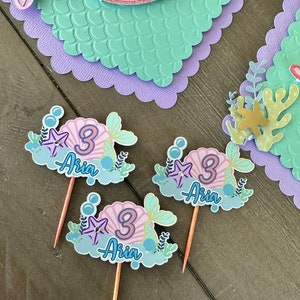Puede incluir: Tres adornos para cupcakes con tem&aacute;tica de sirena con una concha rosa, una estrella de mar dorada y el n&uacute;mero 3. Cada adorno tambi&eacute;n tiene el nombre "Aria" escrito en azul.