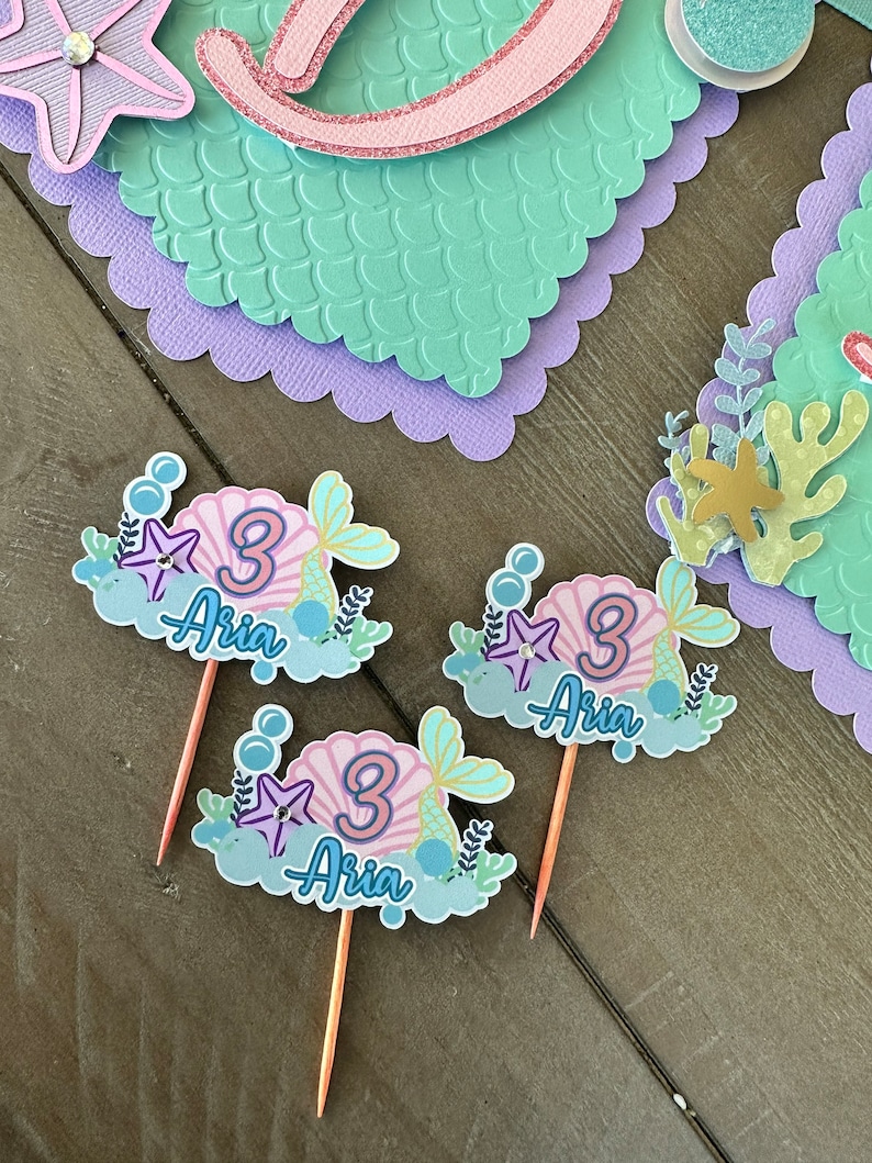Puede incluir: Tres adornos para cupcakes con tem&aacute;tica de sirena con una concha rosa, una estrella de mar dorada y el n&uacute;mero 3. Cada adorno tambi&eacute;n tiene el nombre "Aria" escrito en azul.