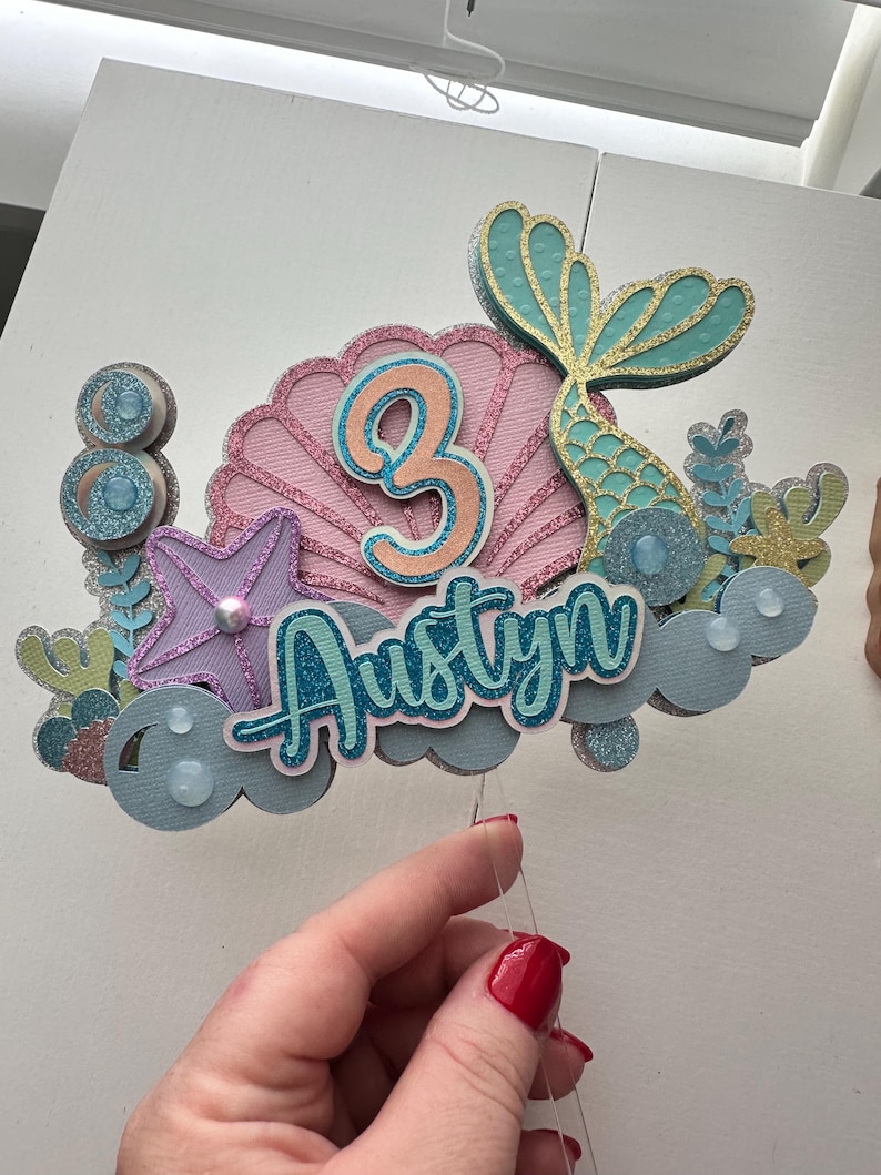 Puede incluir: Un adorno para pastel con tem&aacute;tica de sirena con una concha rosa, una cola de sirena turquesa y el nombre "Austyn" en letras turquesa. El n&uacute;mero "3" est&aacute; en el centro de la concha en color melocot&oacute;n.