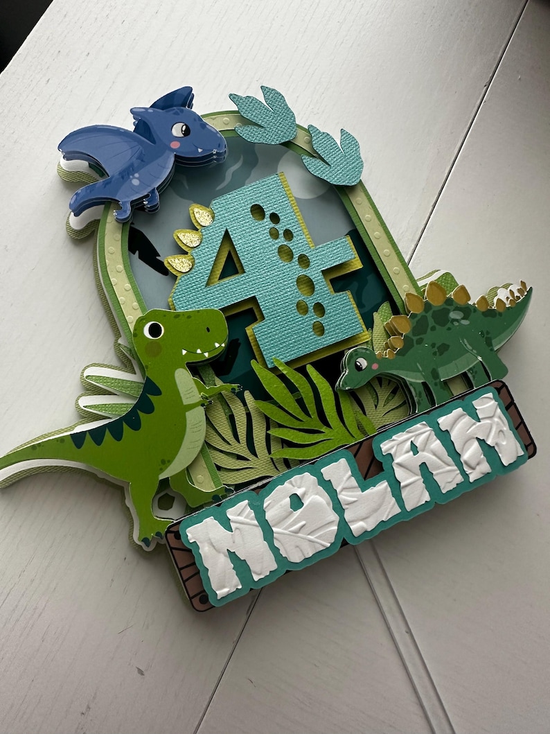 Puede incluir: Un adorno para pastel de dinosaurio verde, azul y amarillo con el n&uacute;mero 4, un pterod&aacute;ctilo y el nombre Nolan.