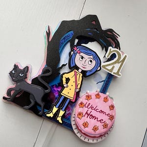 May include: A colorful cake topper featuring a girl with blue hair and a yellow raincoat, a black cat, a hand reaching down from the sky, and a pink cake with the words "Welcome Home" and the number "21".