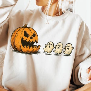 Puede incluir: Una sudadera crema con un gráfico temático de Halloween. El diseño incluye una calabaza y tres fantasmas de dibujos animados. La persona de la imagen sostiene una taza negra. La sudadera es de estilo casual y cómodo.
